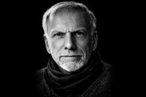 Schwarzweiß-Porträt eines Mannes mit kurzem grauem Haar und grauem Bart, frontal aufgenommen, neutraler Gesichtsausdruck, dunkler Hintergrund.