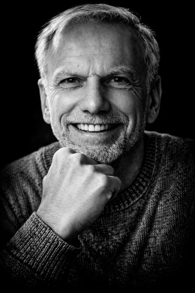 Schwarzweiß-Porträt eines Mannes mit grauem Haar und Bart, Kopf leicht nach vorne geneigt, Hand am Kinn.